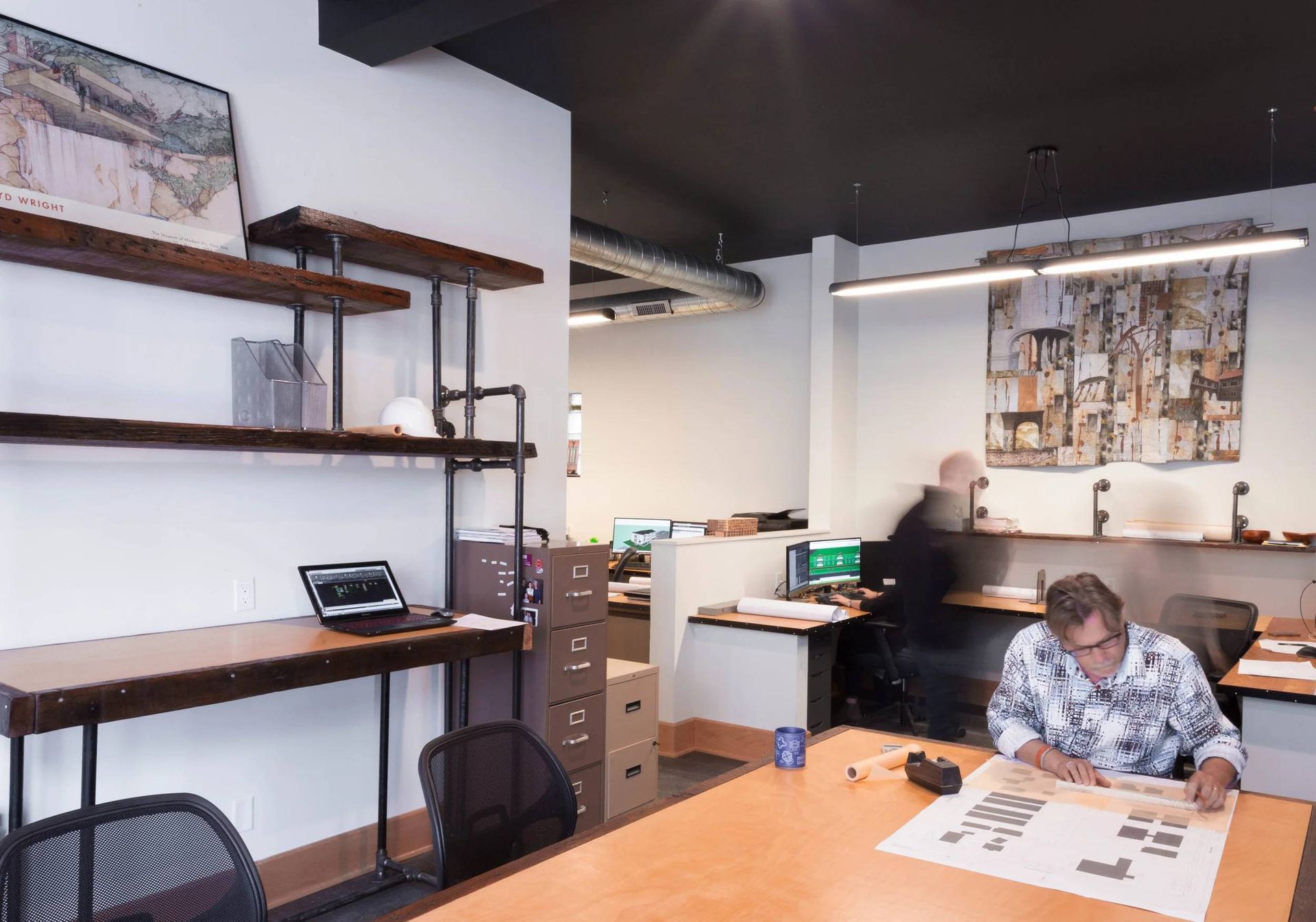 Office with shelves and desks; a person sits at a table reviewing papers in a bright workspace.