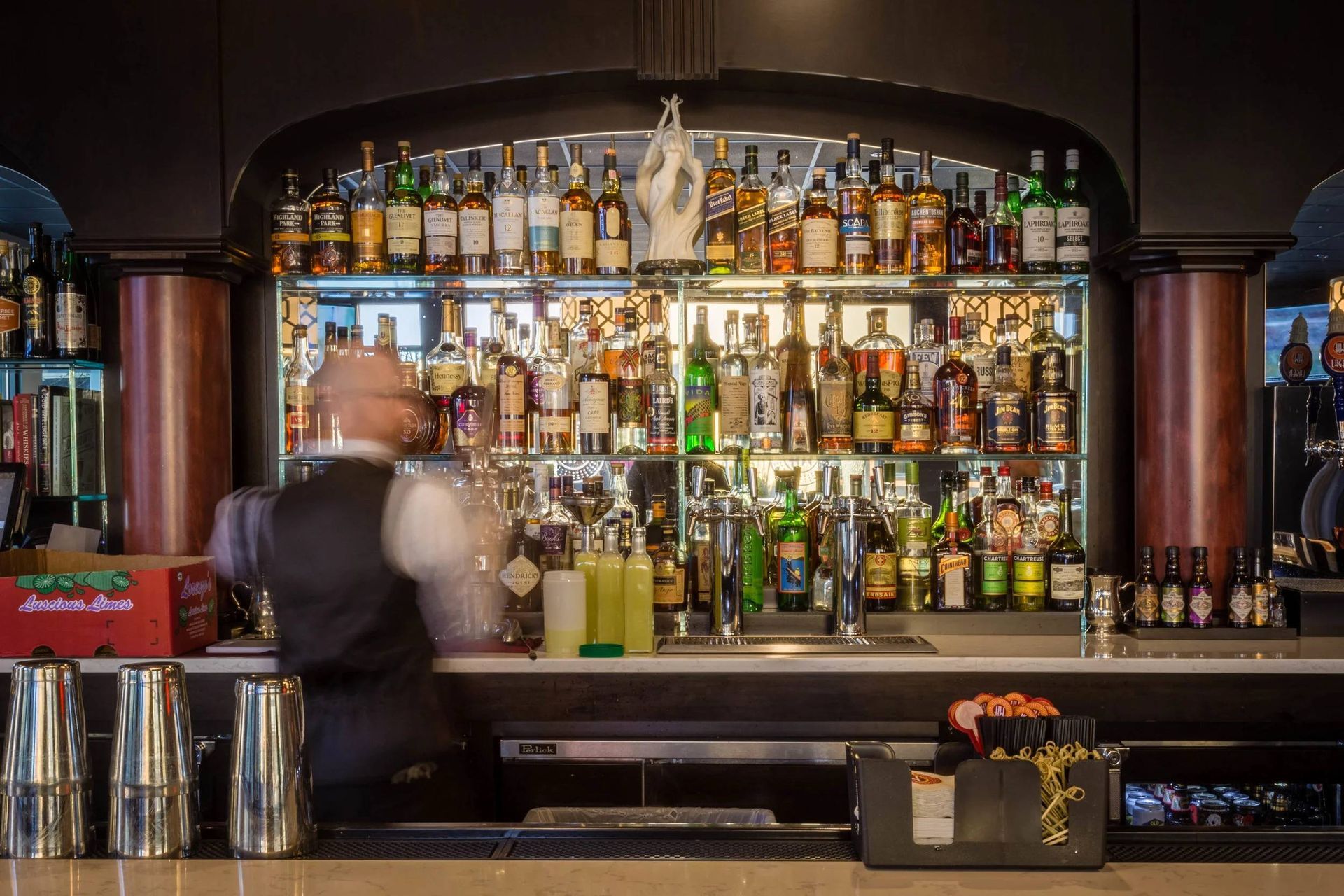 Blurred bartender behind a stocked liquor bar with rows of bottles and glass shelves