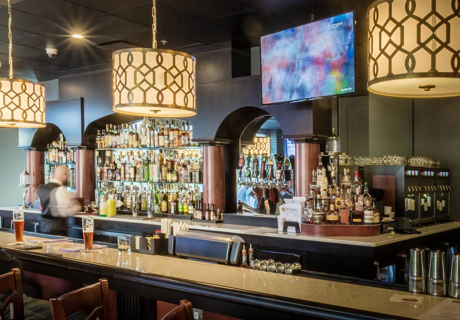 Modern bar interior with illuminated shelves, hanging lamps, and a colorful TV screen