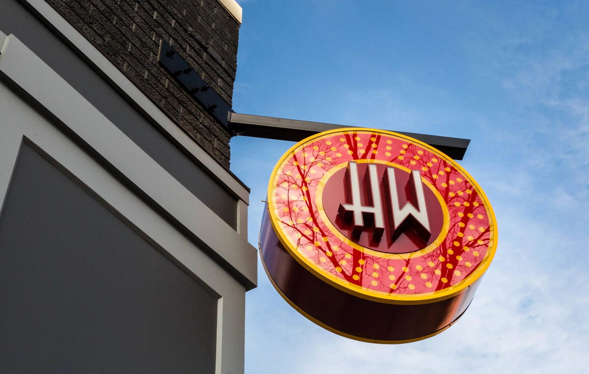 A round red and yellow sign with a stylized black logo hangs on a building against a blue sky.