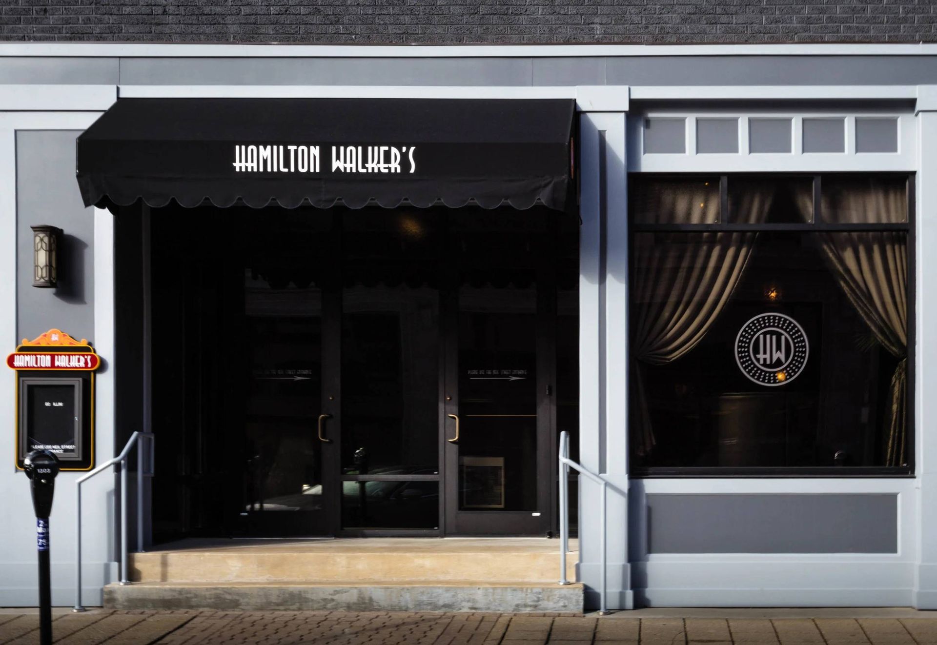 Storefront with black awning, dark glass entrance, and a window display with curtains and a circular logo.