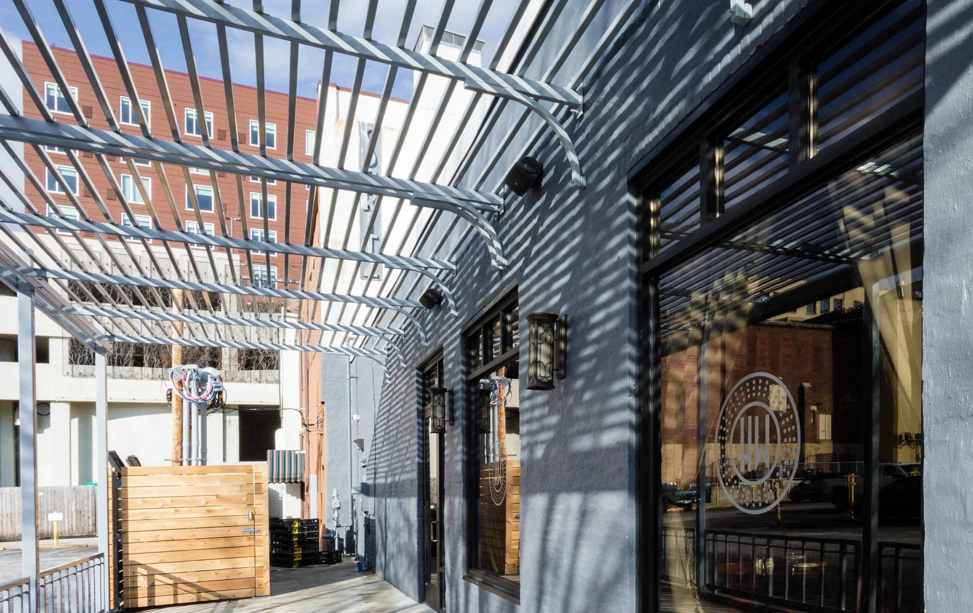 Sunlit outdoor patio with metal pergola, brick building, and glass doors with striped shadows