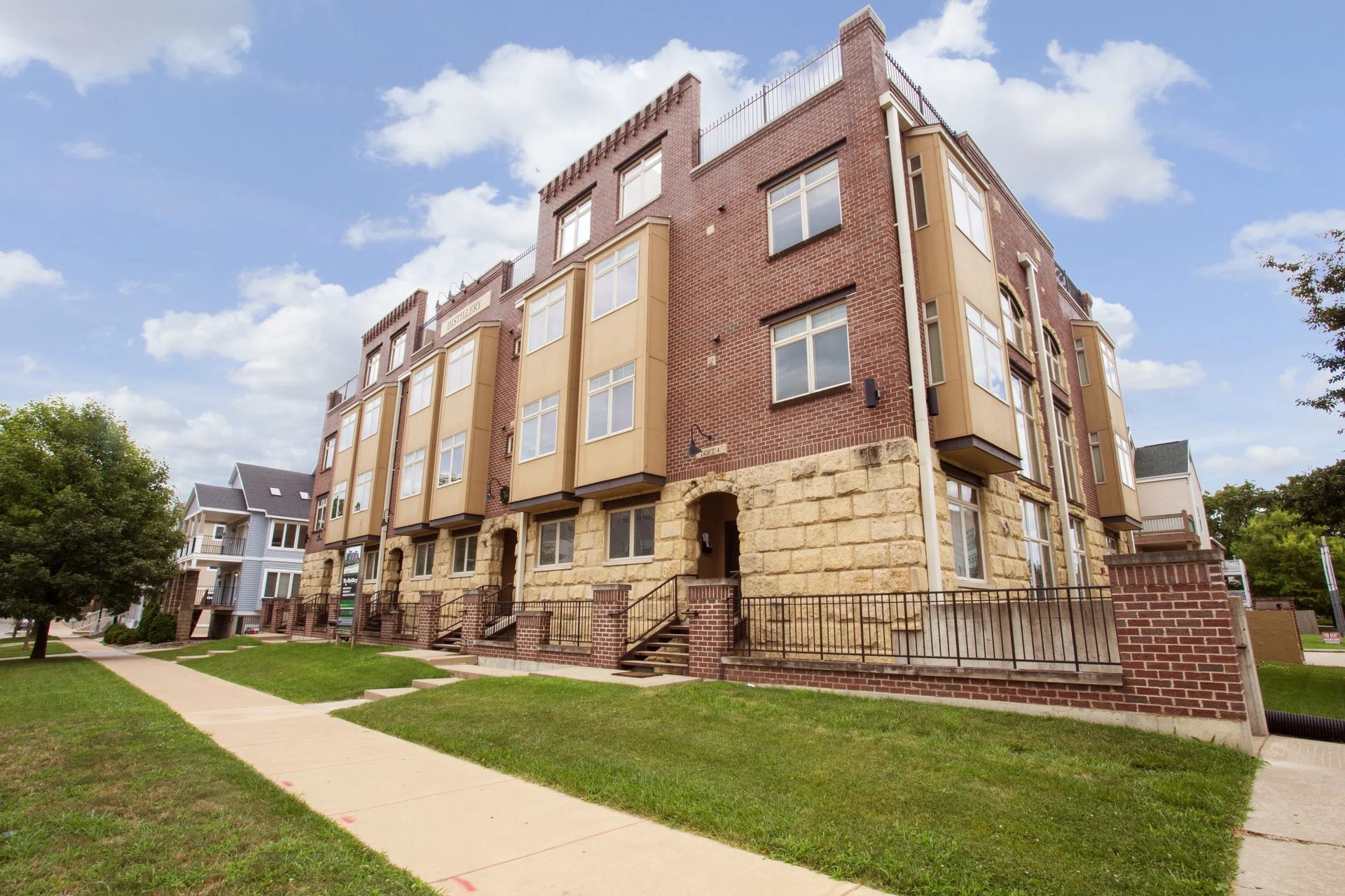 Brick apartment building with tan stone lower facade beside a sidewalk and grassy lawn under a partly cloudy sky