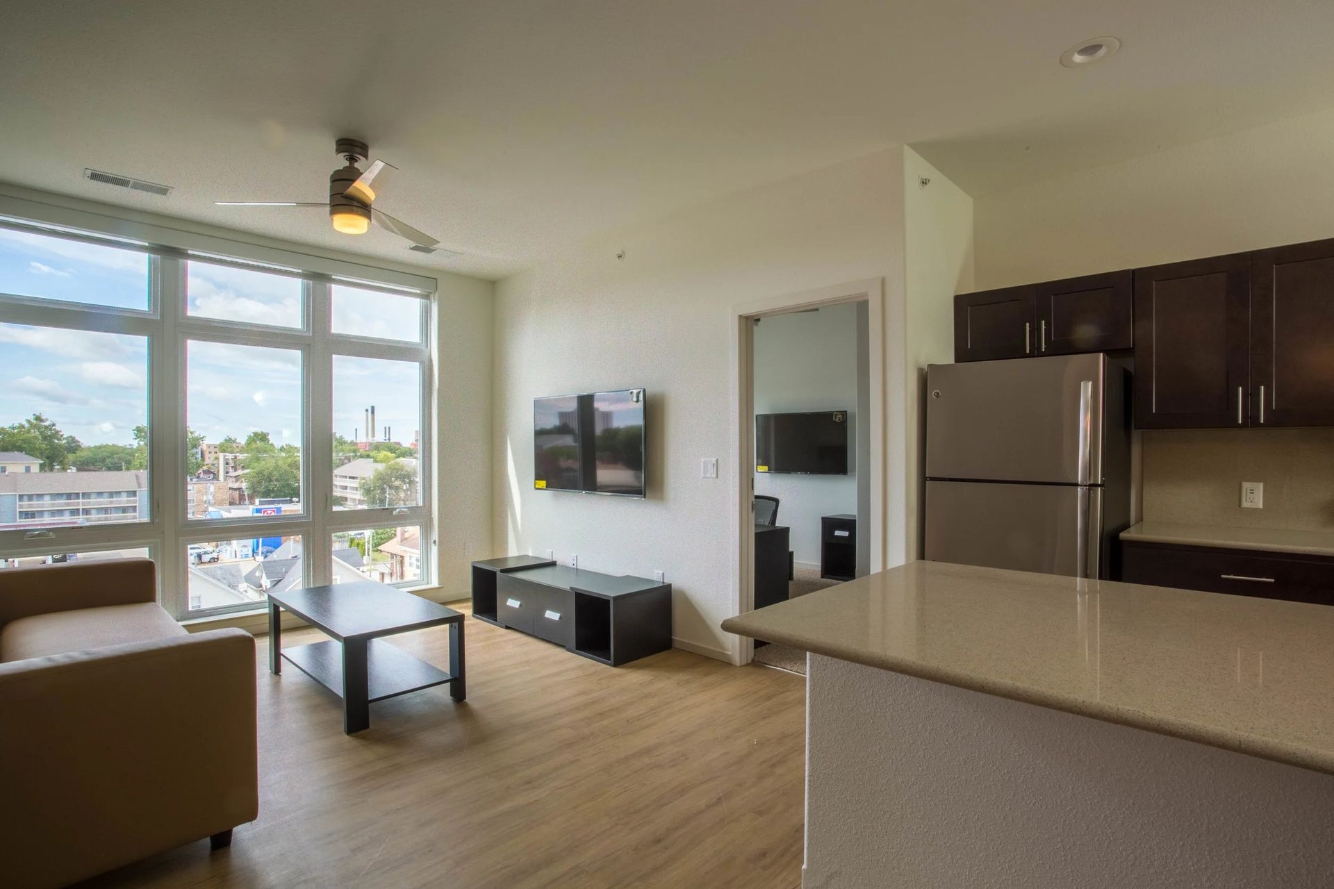 Bright open living room with kitchen, large windows, sofa, TV, and city view