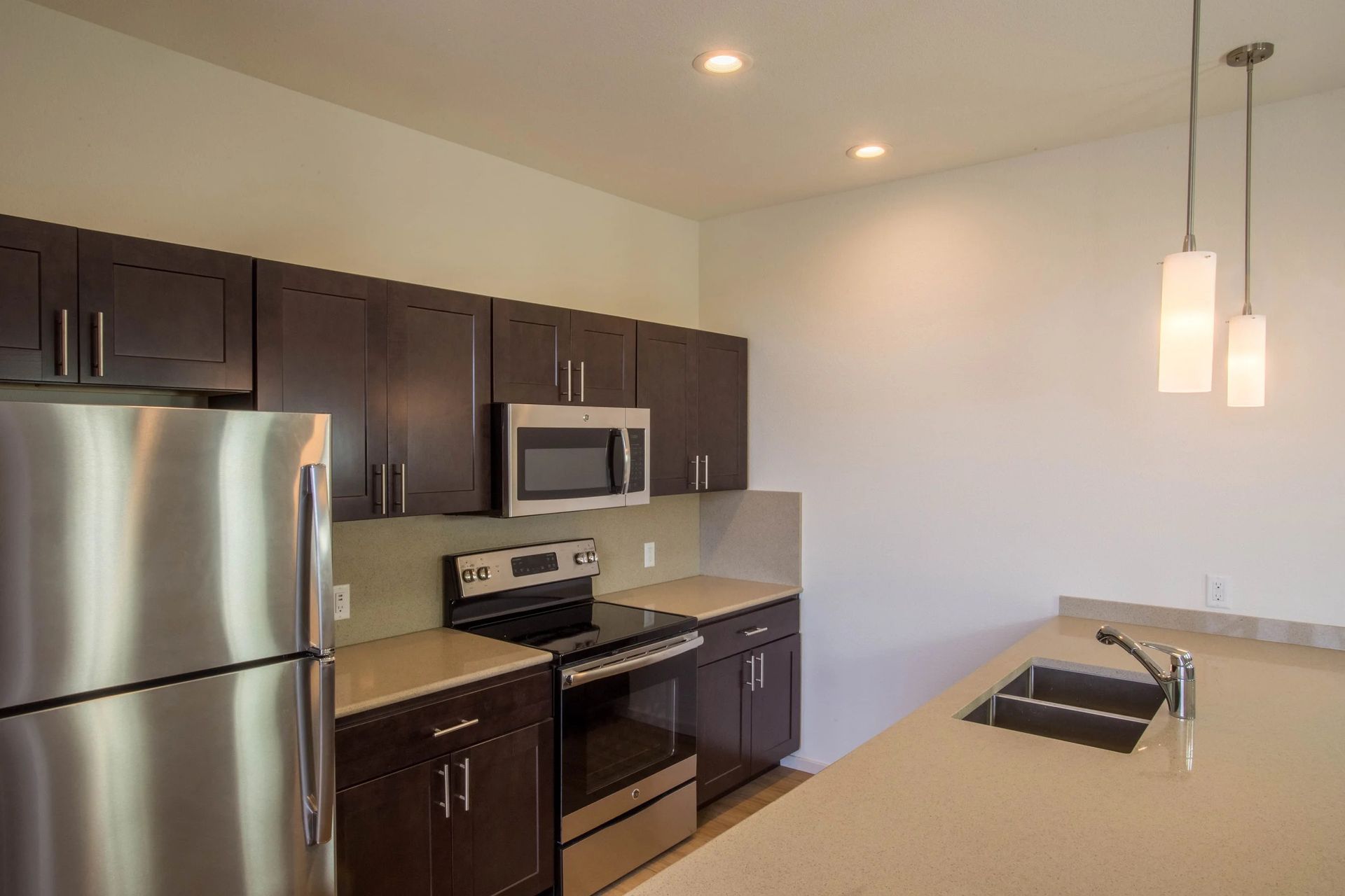 Modern kitchen with stainless steel appliances, dark cabinets, beige counters, and a breakfast bar with pendant lights