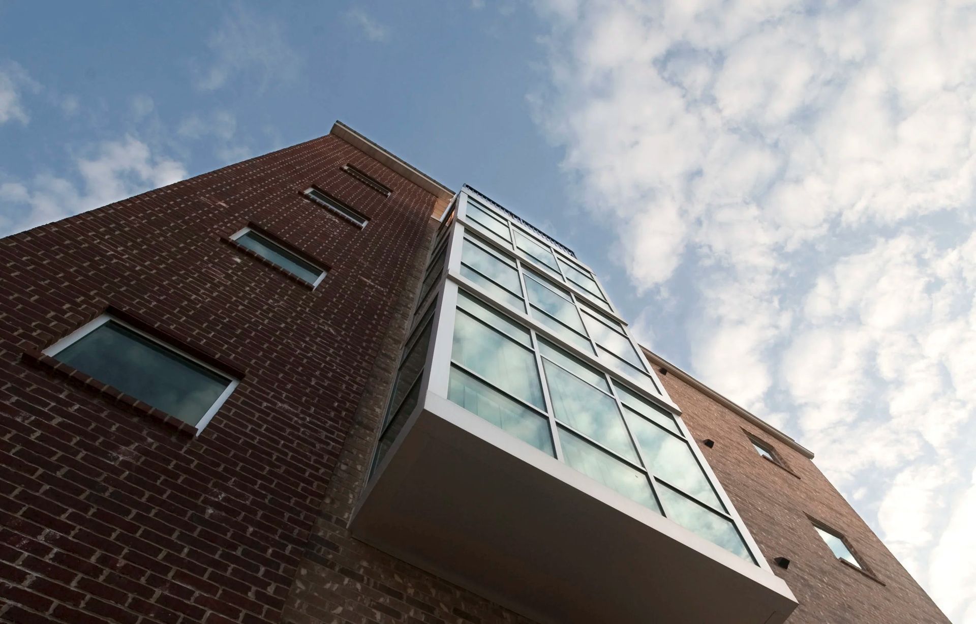Low-angle view of a brick apartment building with a glass balcony against a cloudy sky