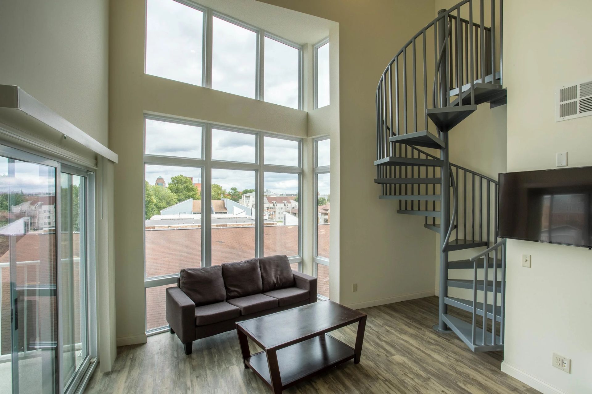 Bright living room with large windows, brown sofa, coffee table, and spiral staircase.