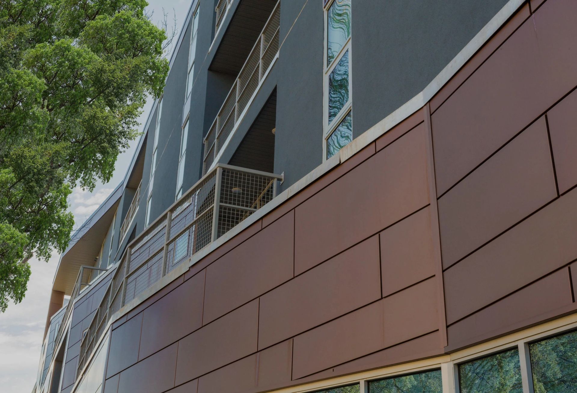Modern building facade with brown panels, blue-gray upper walls, and trees beside it
