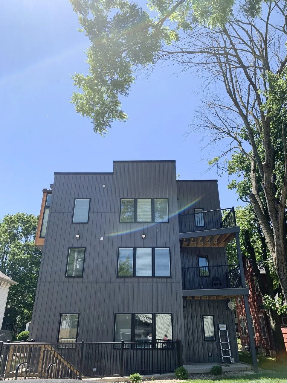 Modern dark-gray apartment building with balconies under a clear blue sky, framed by trees
