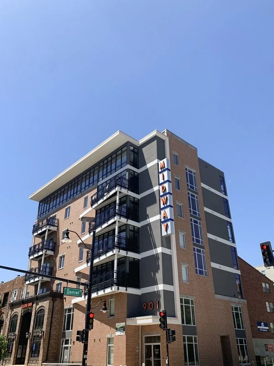 Modern multi-story brick building with balconies and a rooftop overhang under a clear blue sky