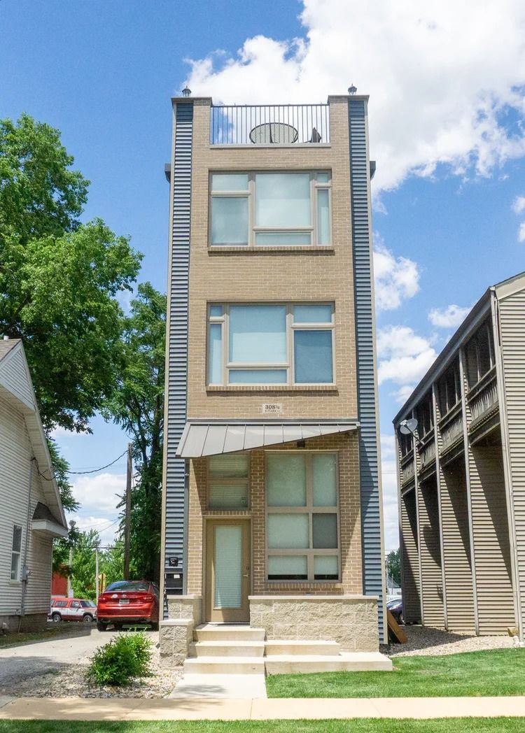 Three-story tan brick house with a rooftop railing, large front windows, and a small porch under a blue sky.