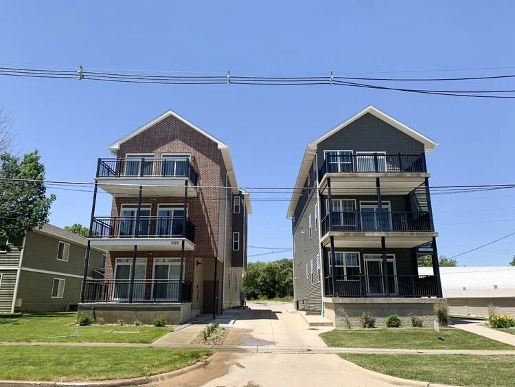 Two three-story apartment buildings with balconies on a sunny street, separated by a driveway