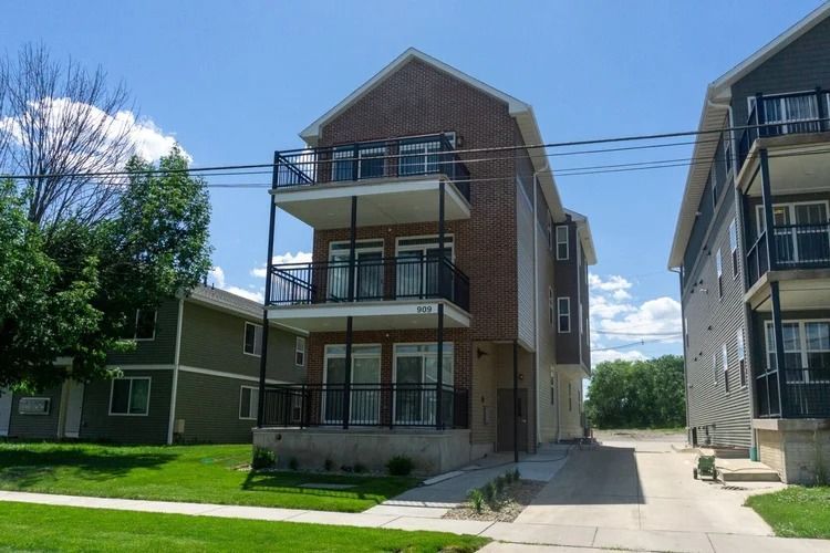 Three-story apartment building with balconies beside a walkway and green lawn under a blue sky