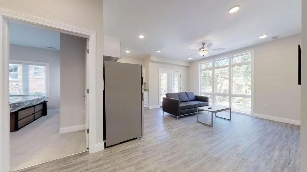 Bright open living room with large windows, gray sofa, glass coffee table, and adjoining kitchen area