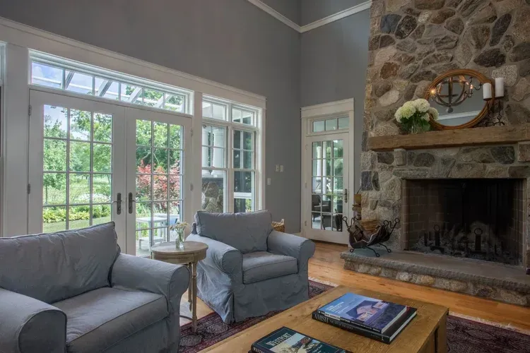 Bright living room with gray chairs, stone fireplace, and large windows overlooking greenery