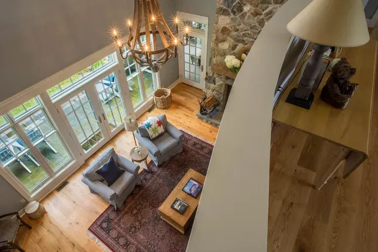 Bright living room with two armchairs, rug, fireplace, chandelier, and tall windows overlooking a wooded view