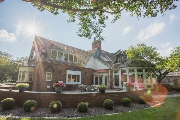 Sunlit brick cottage with white porch, landscaped shrubs, and trees in front yard