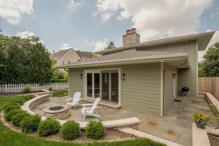 Backyard patio with fire pit, white chairs, and sliding glass doors beside a light gray house