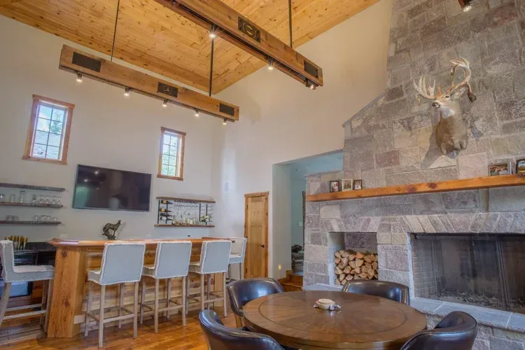 Modern living room with wood beams, dining table, wall TV, and large stone fireplace