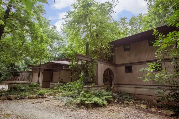 Brown house nestled in dense green trees with a stone path and arched front door
