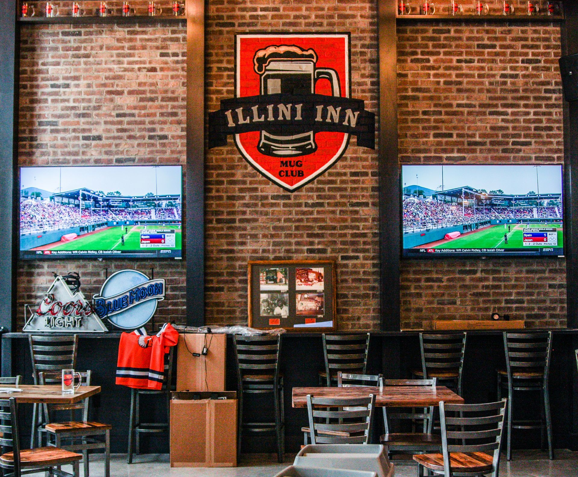 Brick sports bar interior with Illini Inn sign, two TVs, high tables, and chairs