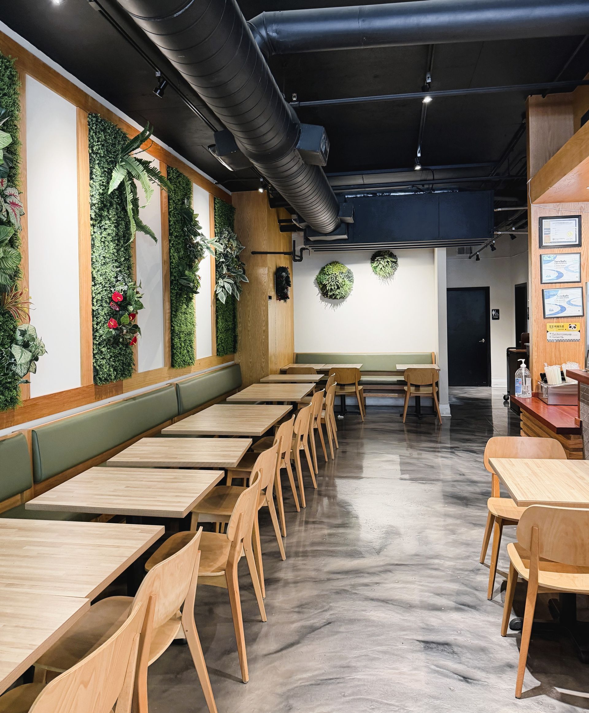 Modern café interior with wooden tables and chairs, wall greenery, and exposed black ceiling ducts.
