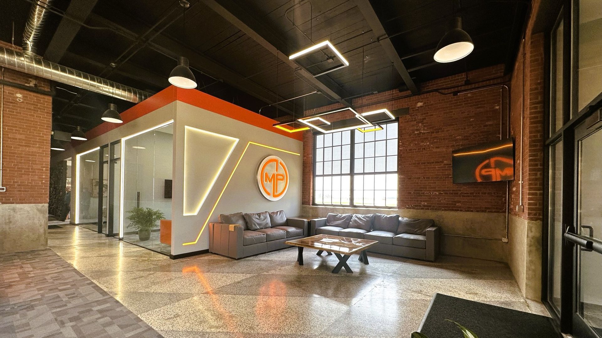 Modern office lounge with brick walls, glass pod, orange accents, and seating under industrial lighting