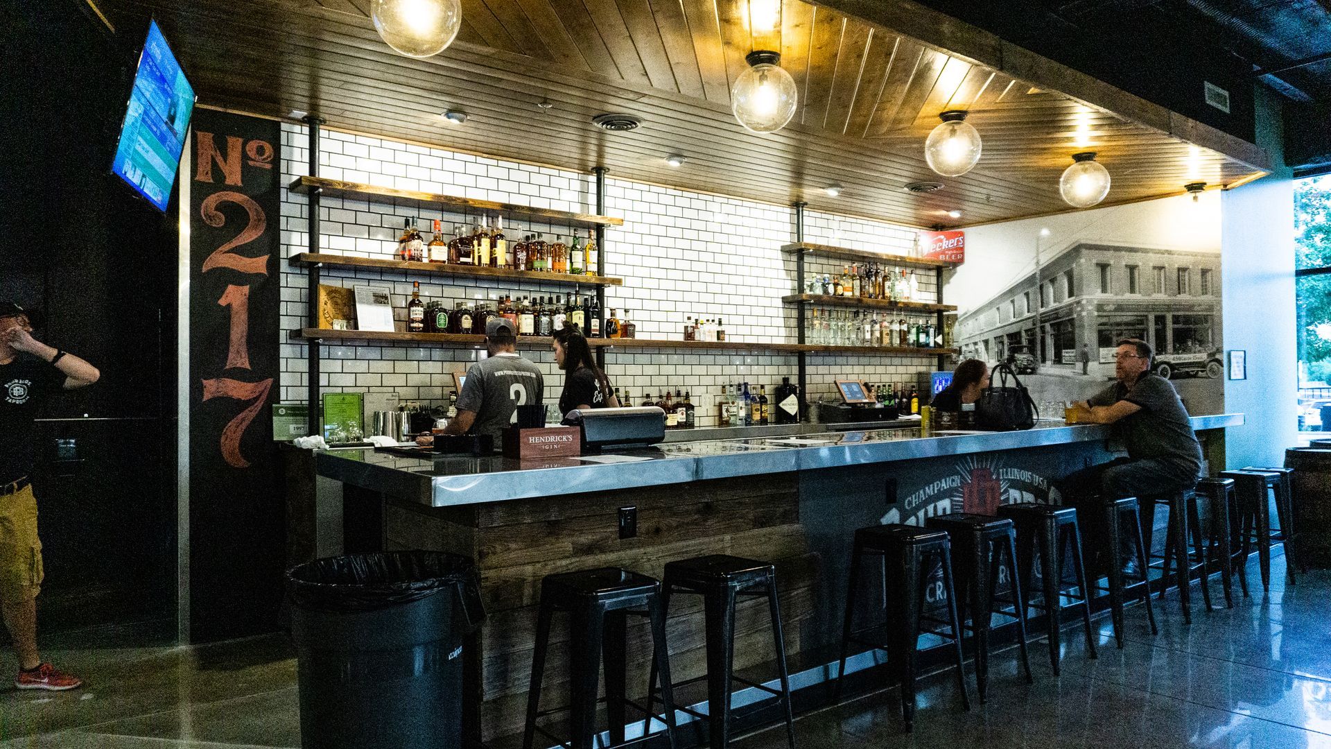 Dim bar interior with a long counter, stools, pendant lights, and shelves of bottles behind the bar.