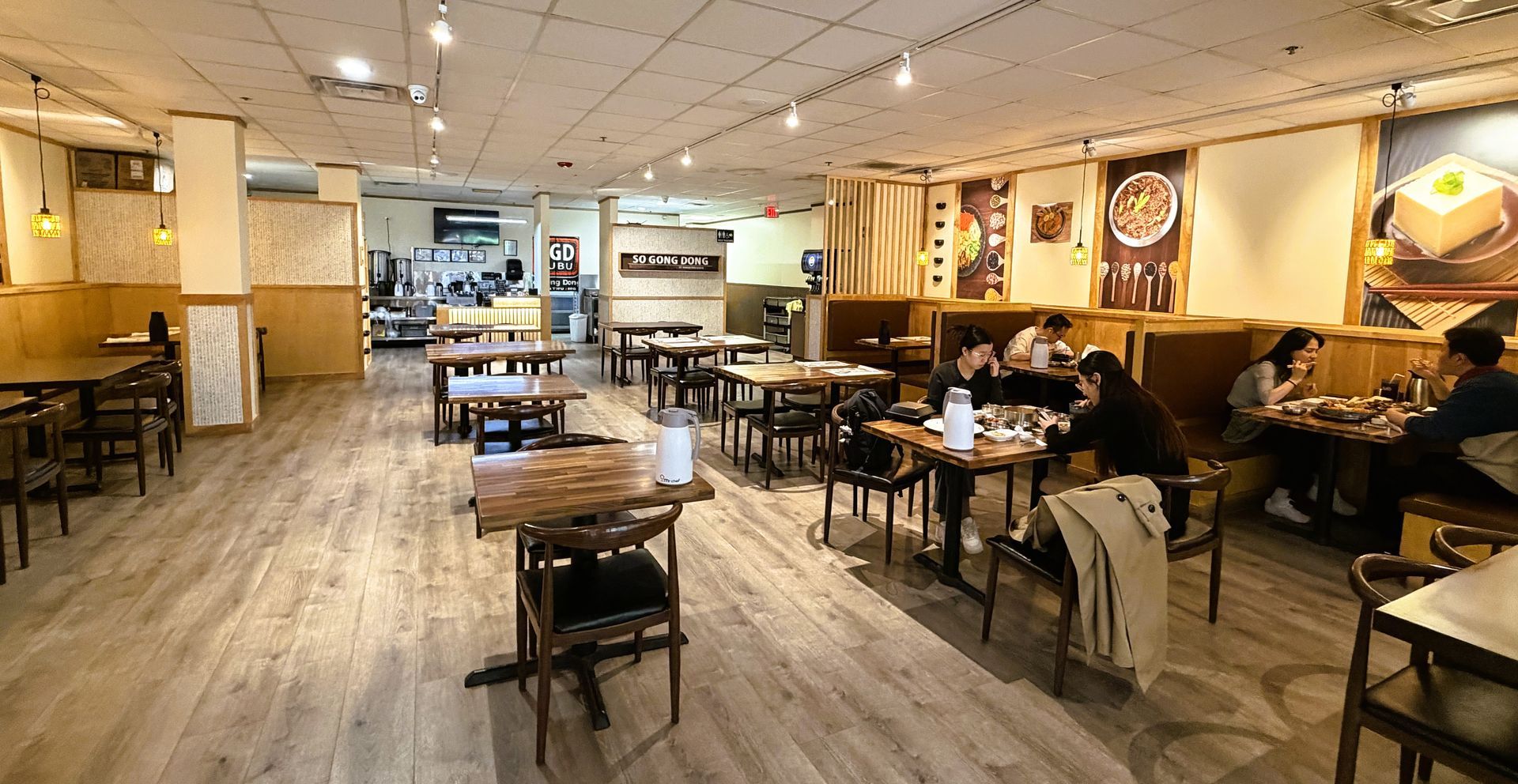Cozy café interior with wooden tables, warm lighting, wall art, and a few customers seated at the right.