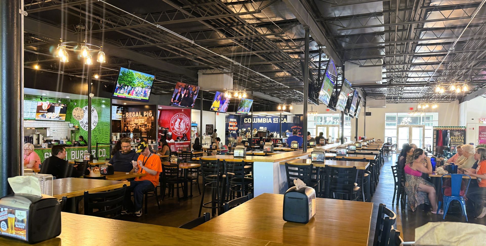 Spacious indoor food hall with tables, hanging lights, TVs, and people dining