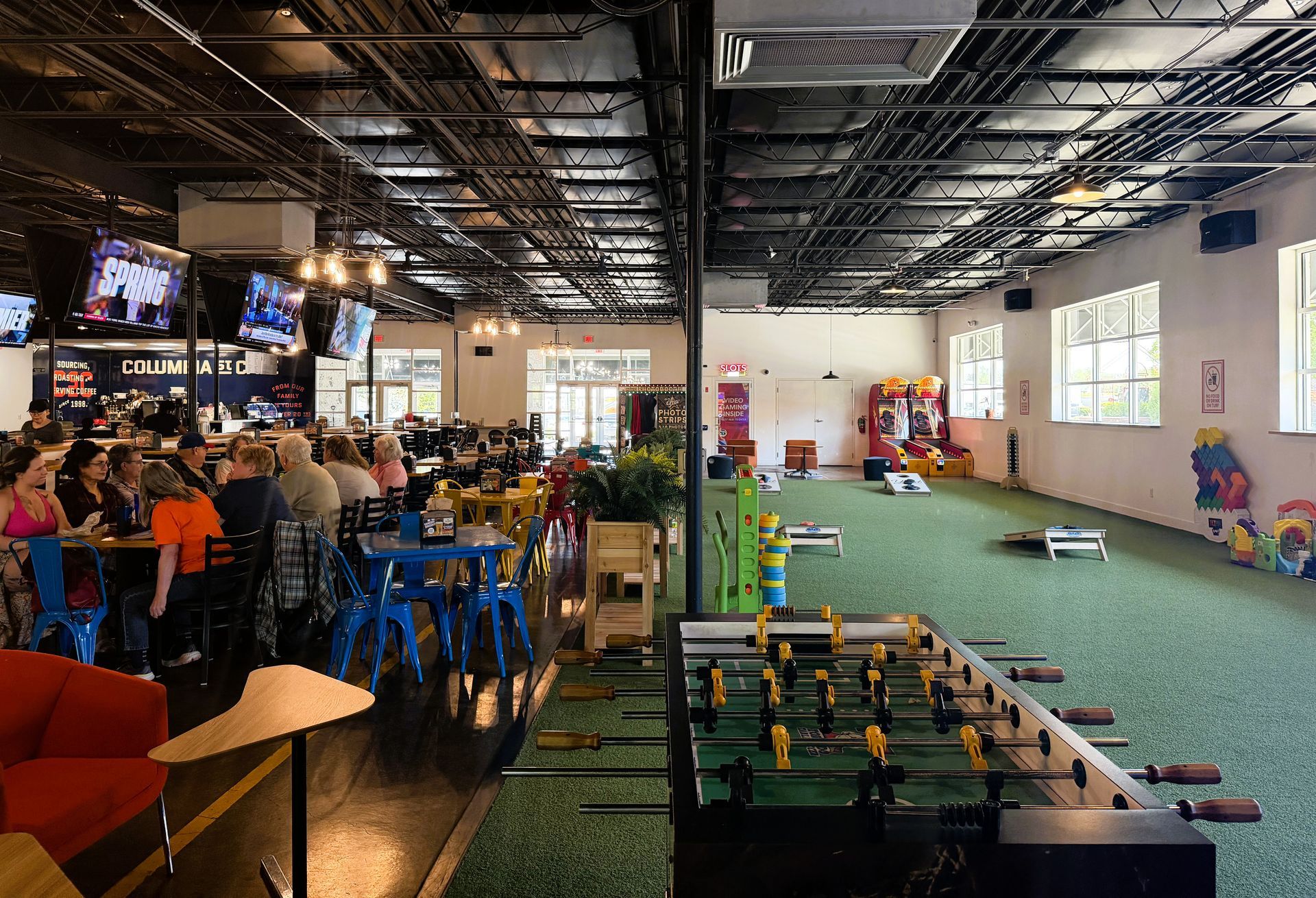 Indoor mini-golf and arcade area with green turf, colorful arcade games, and people seated at tables