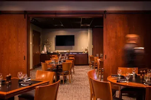 Elegant restaurant dining room with wooden tables, warm lighting, and a blurred server walking by