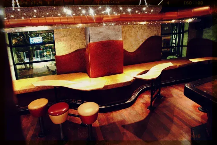 Empty nightclub interior with curved red-and-gold bar, stools, and wood flooring under warm lights