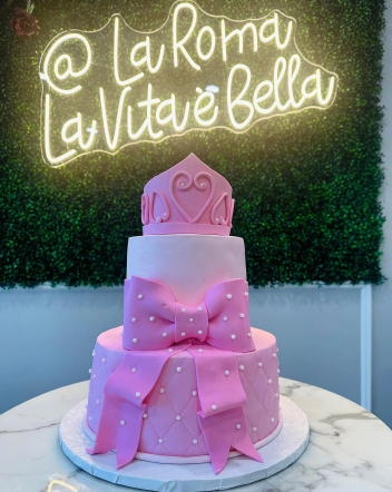 A pink cake with a bow and pearls is on a table in front of a neon sign that says la roma la vita e bella