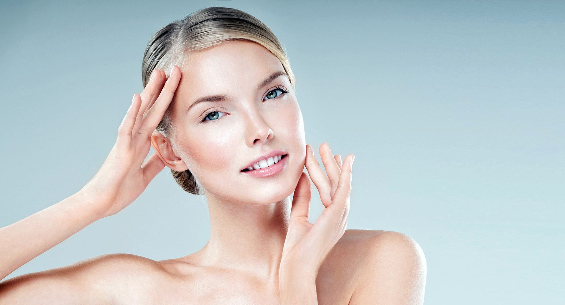 Woman with fair skin touches her face, smiling. Light blue background.