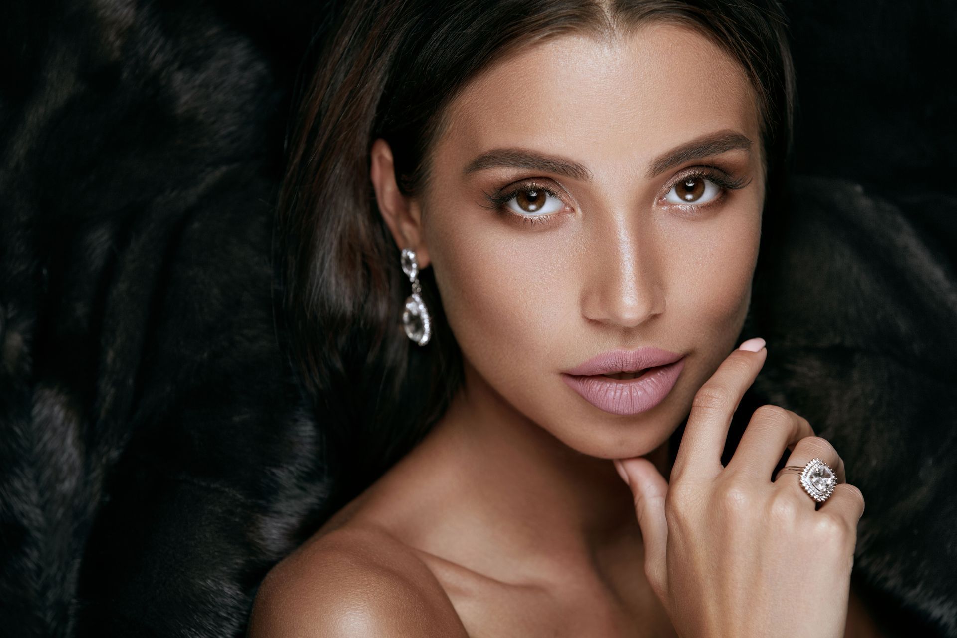 Woman with dark hair, wearing earrings and a ring, poses with a hand near her face, dark background.