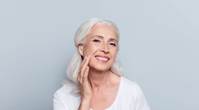 Smiling woman with short white hair touching her cheek against a light gray background