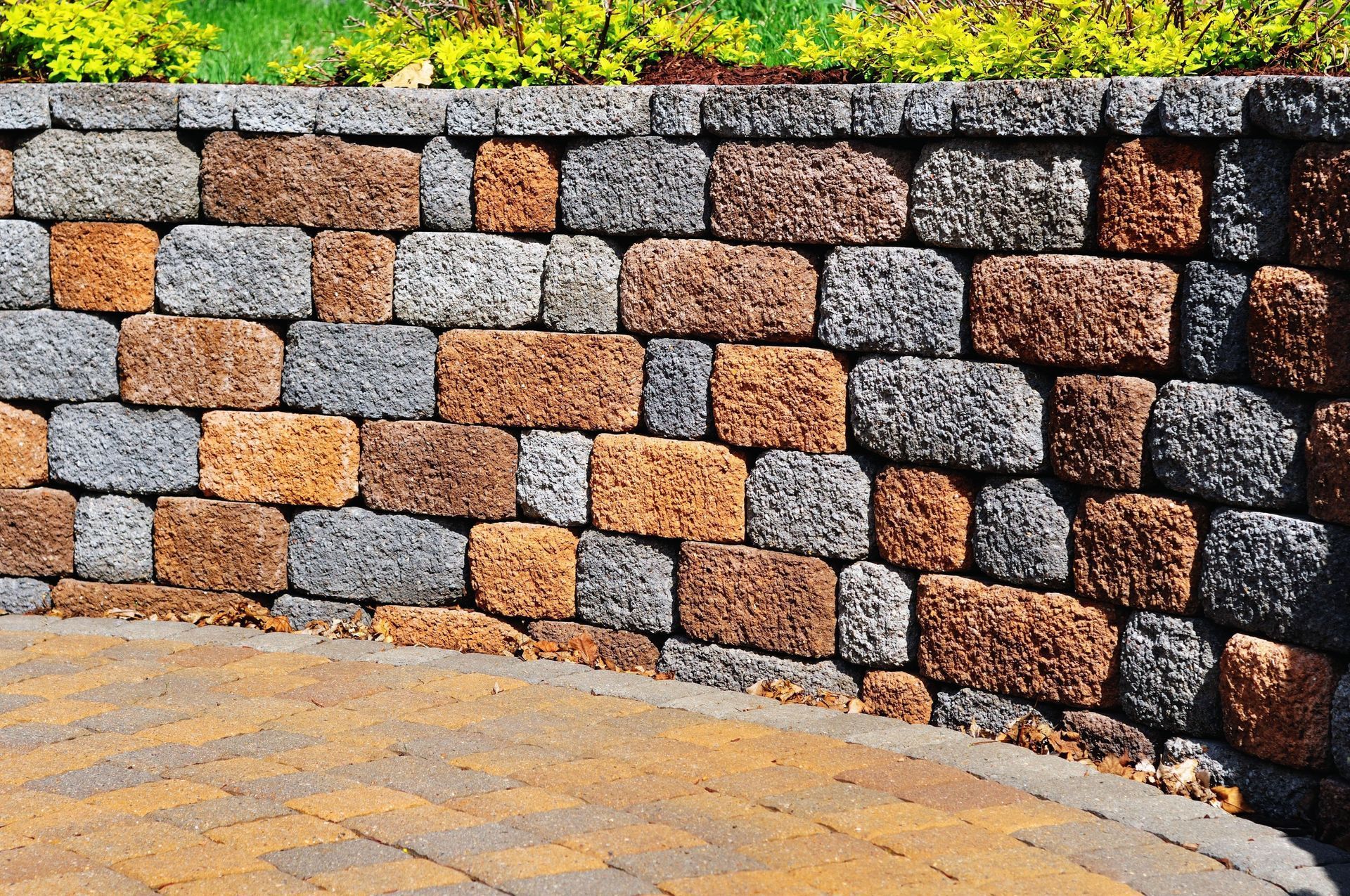 Stone retaining wall made of orange and gray blocks, curved edge, pavers below, greenery above.