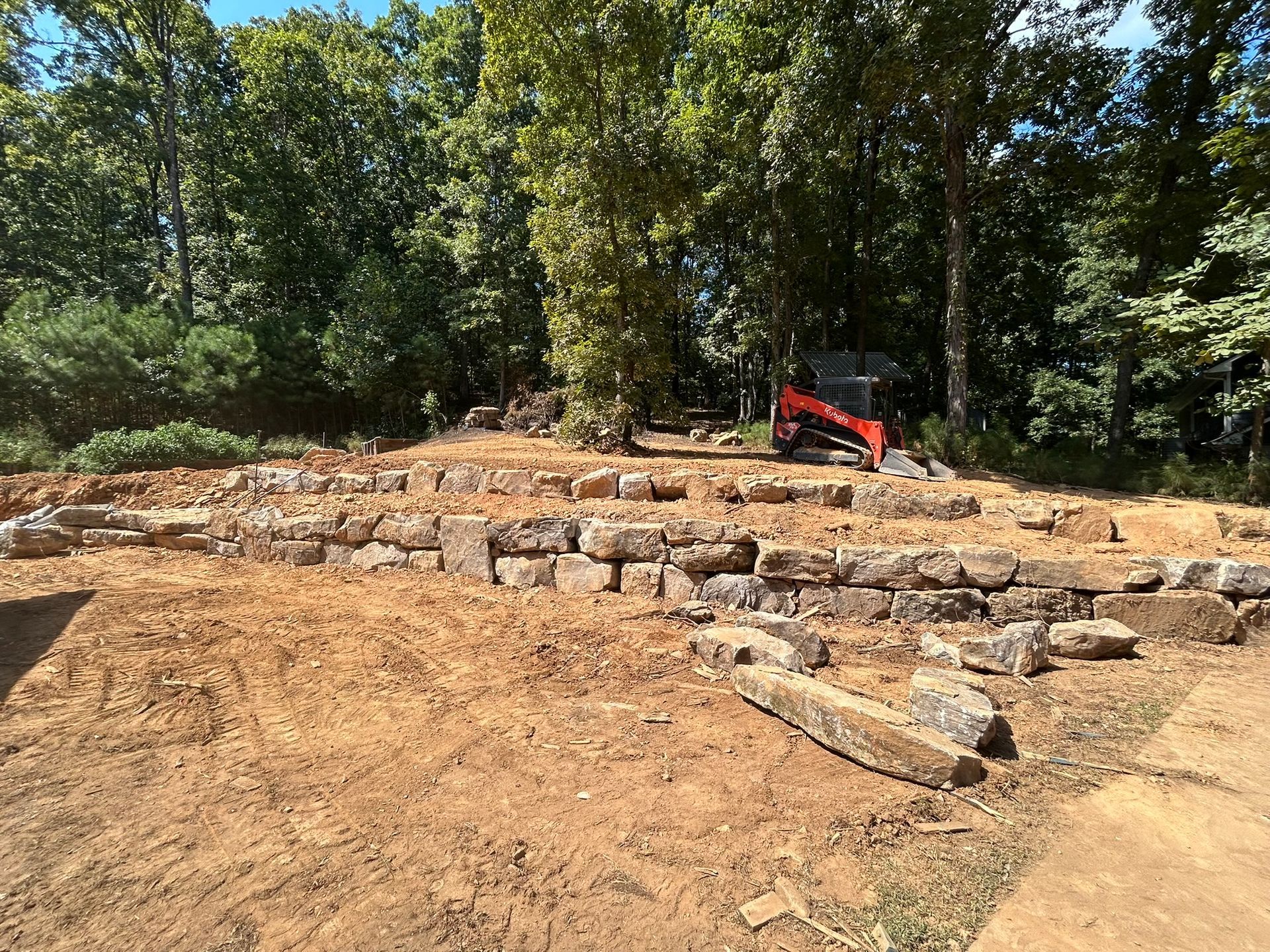 Stone retaining walls and dirt cleared area with a small excavator in the background, trees in the background.
