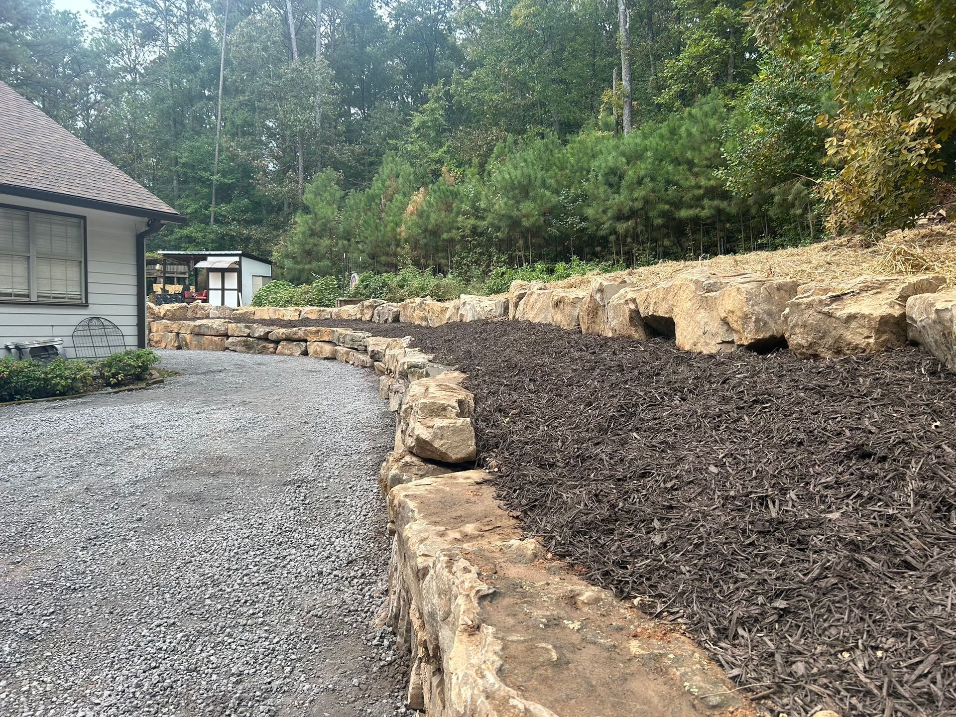 Gravel driveway next to a stone retaining wall filled with dark mulch; trees in the background.