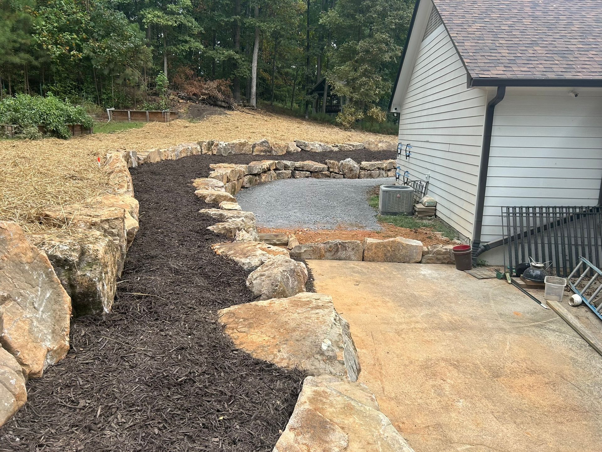 Landscape with a rock retaining wall, mulch, and a concrete patio next to a house.