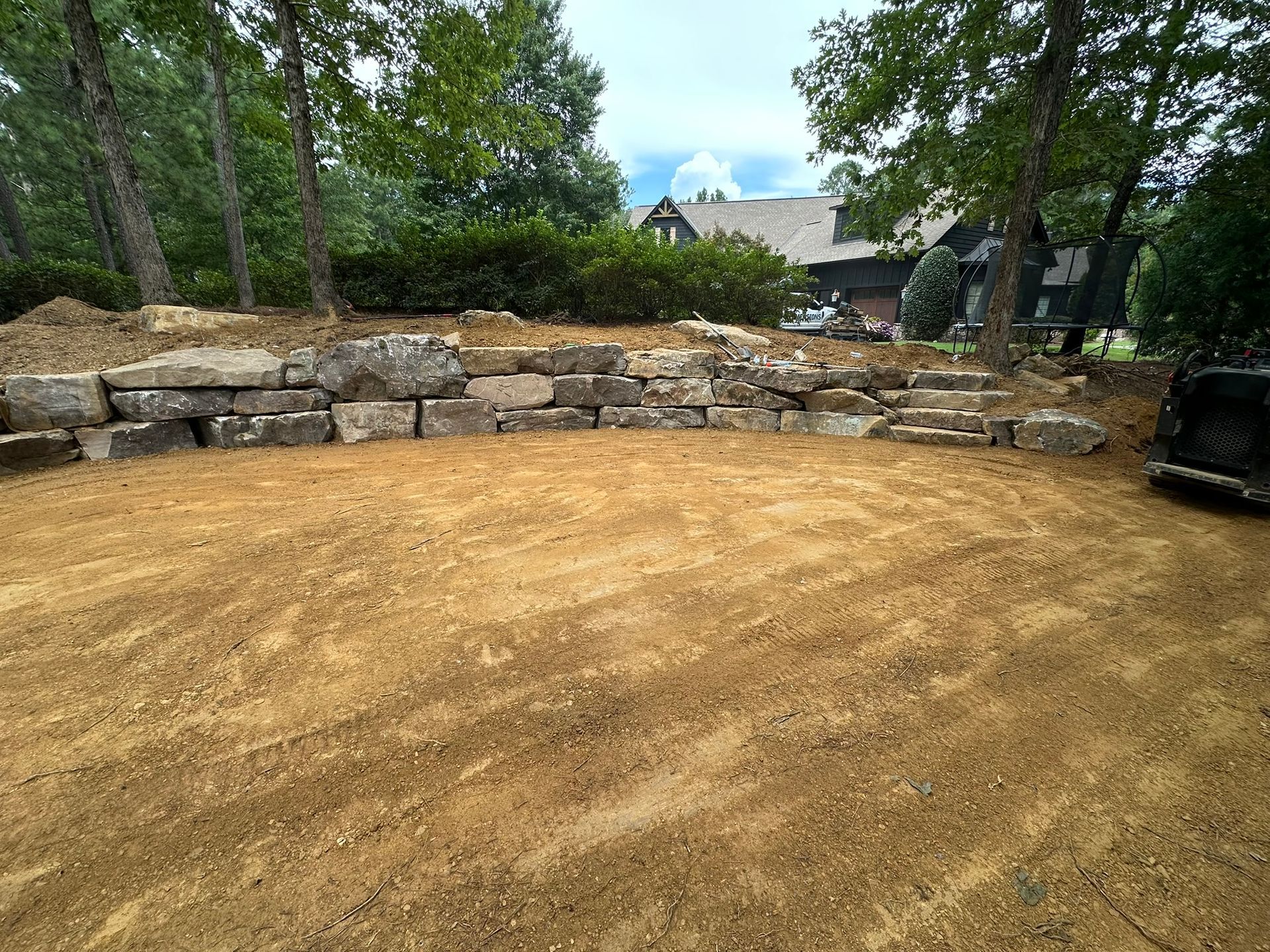 A dirt area in front of a stone retaining wall with trees and a house in the background.