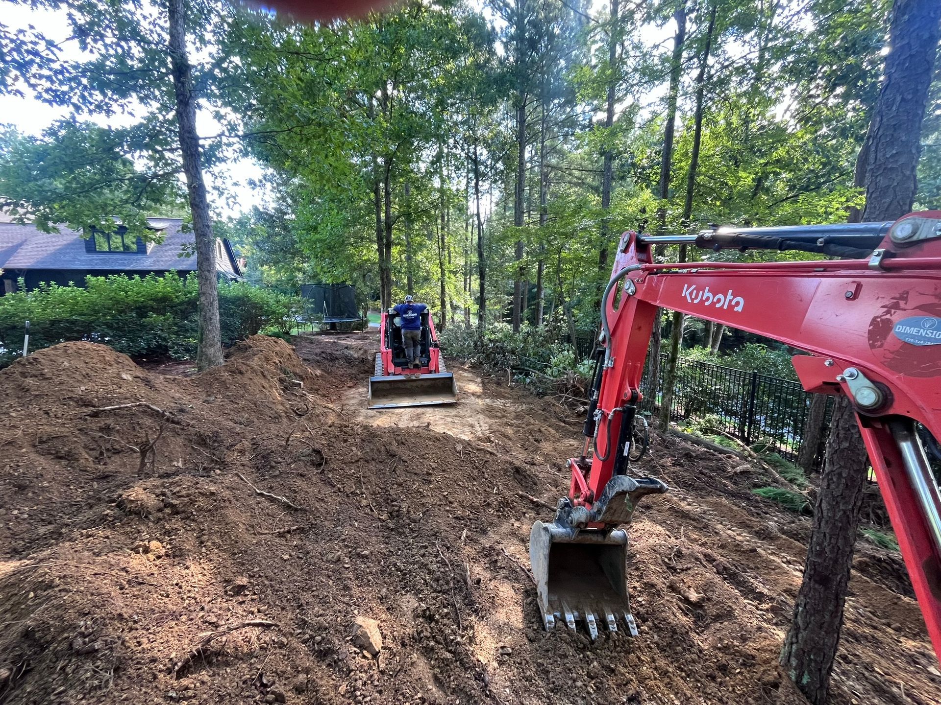 Excavator and mini-excavator digging in a wooded area; dirt mound in foreground, trees and house in background.