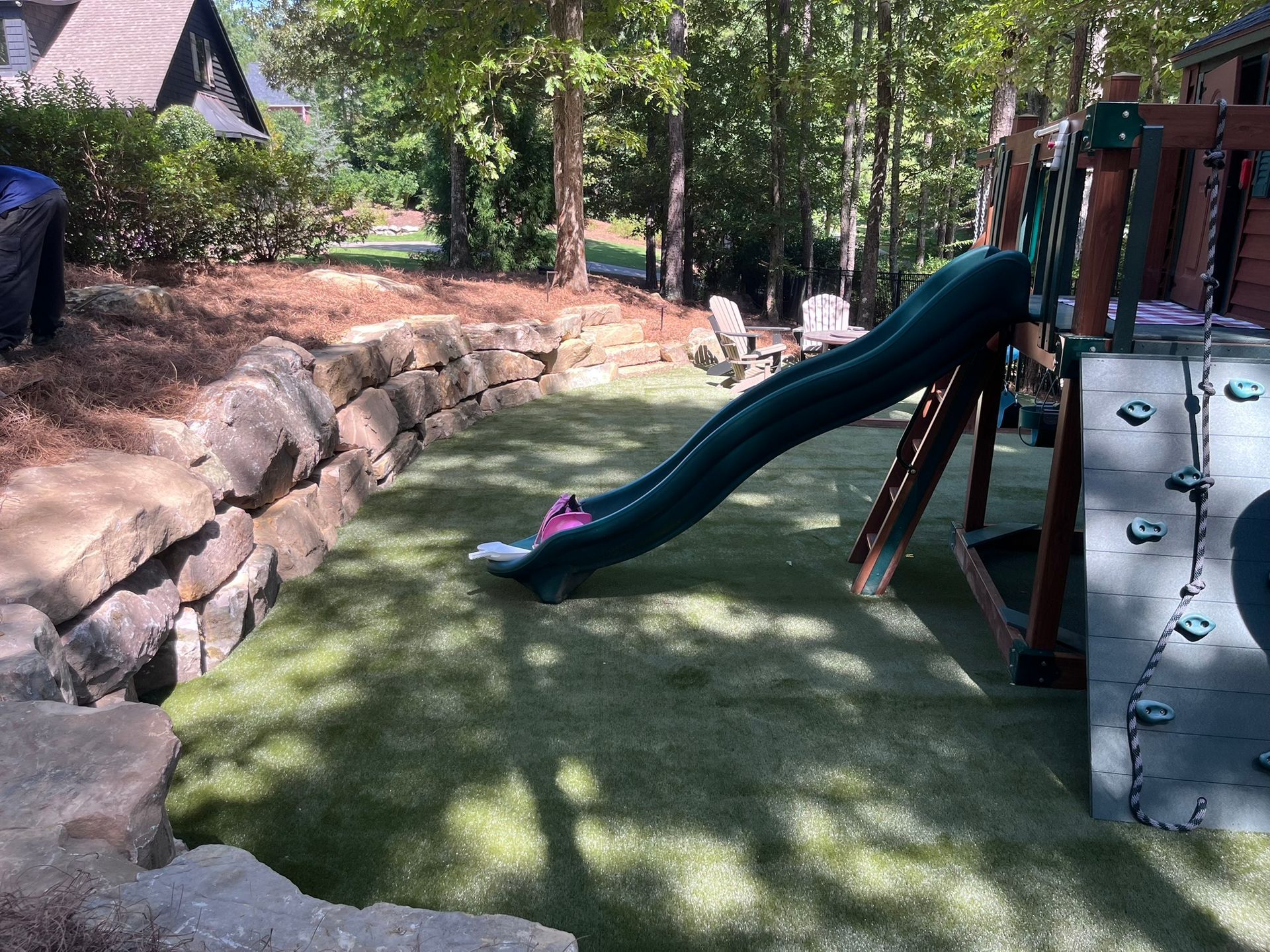 Playground with green slide and climbing wall on artificial turf, next to a stone retaining wall.