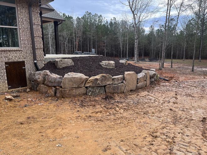 Stone retaining wall holding mulch bed next to a brick house. Wooded background.