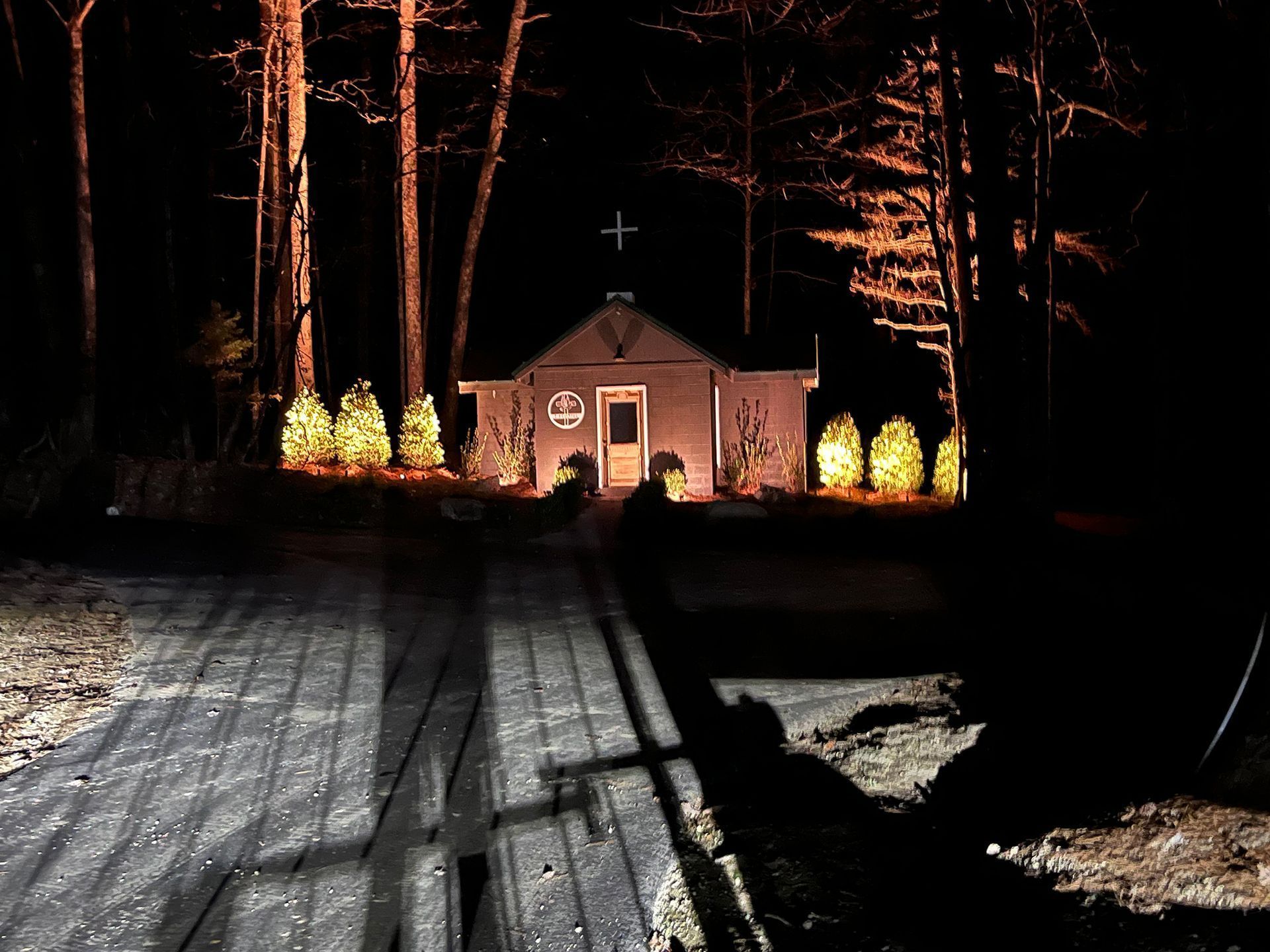 Small chapel at night, illuminated, with a cross atop. Trees line the pathway.