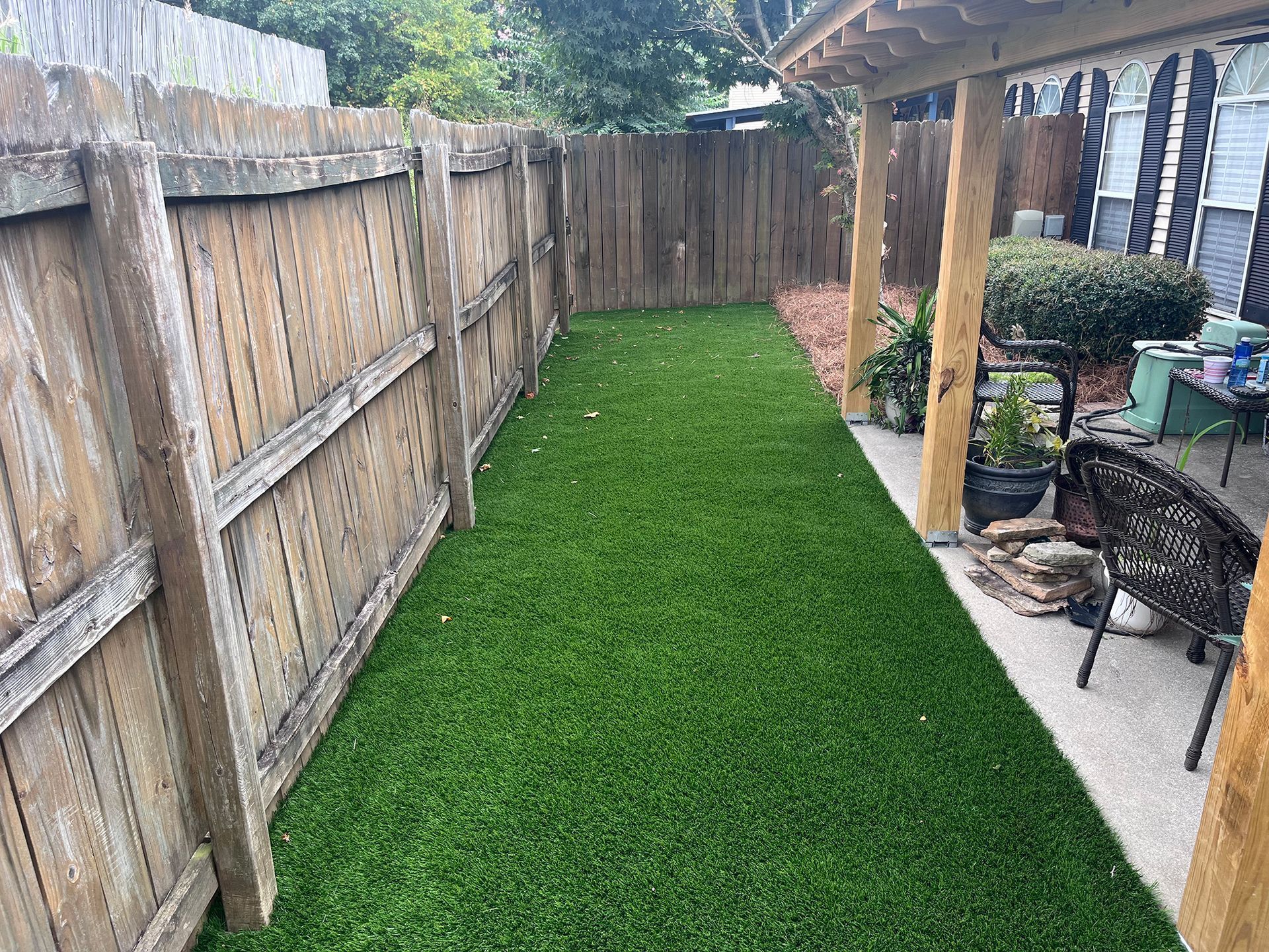 Narrow backyard with artificial turf, wooden fence, and covered patio with plants.