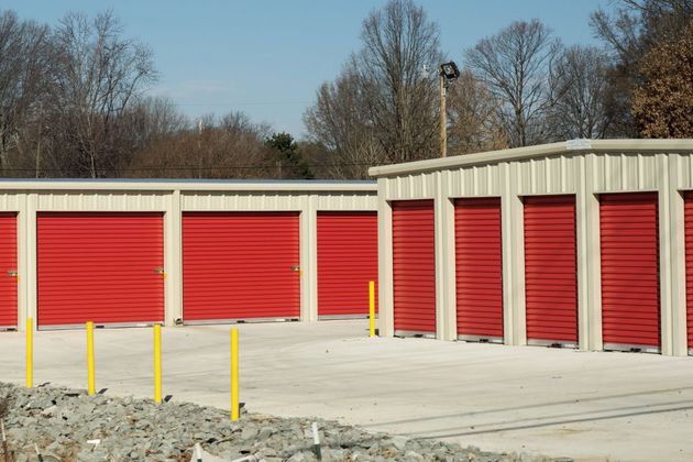 A row of storage units with red metal roll-up doors against a background of bare trees under a clear blue sky.