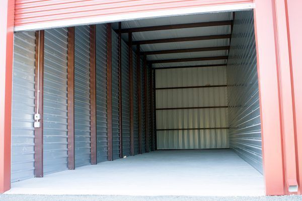 An empty commercial storage unit with a red door frame, gray corrugated metal walls, and concrete flooring.