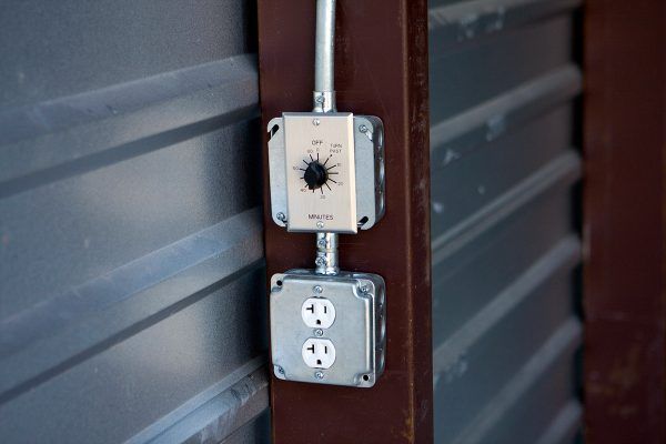 An electrical control knob and a dual power outlet mounted on a metal post against a corrugated wall.